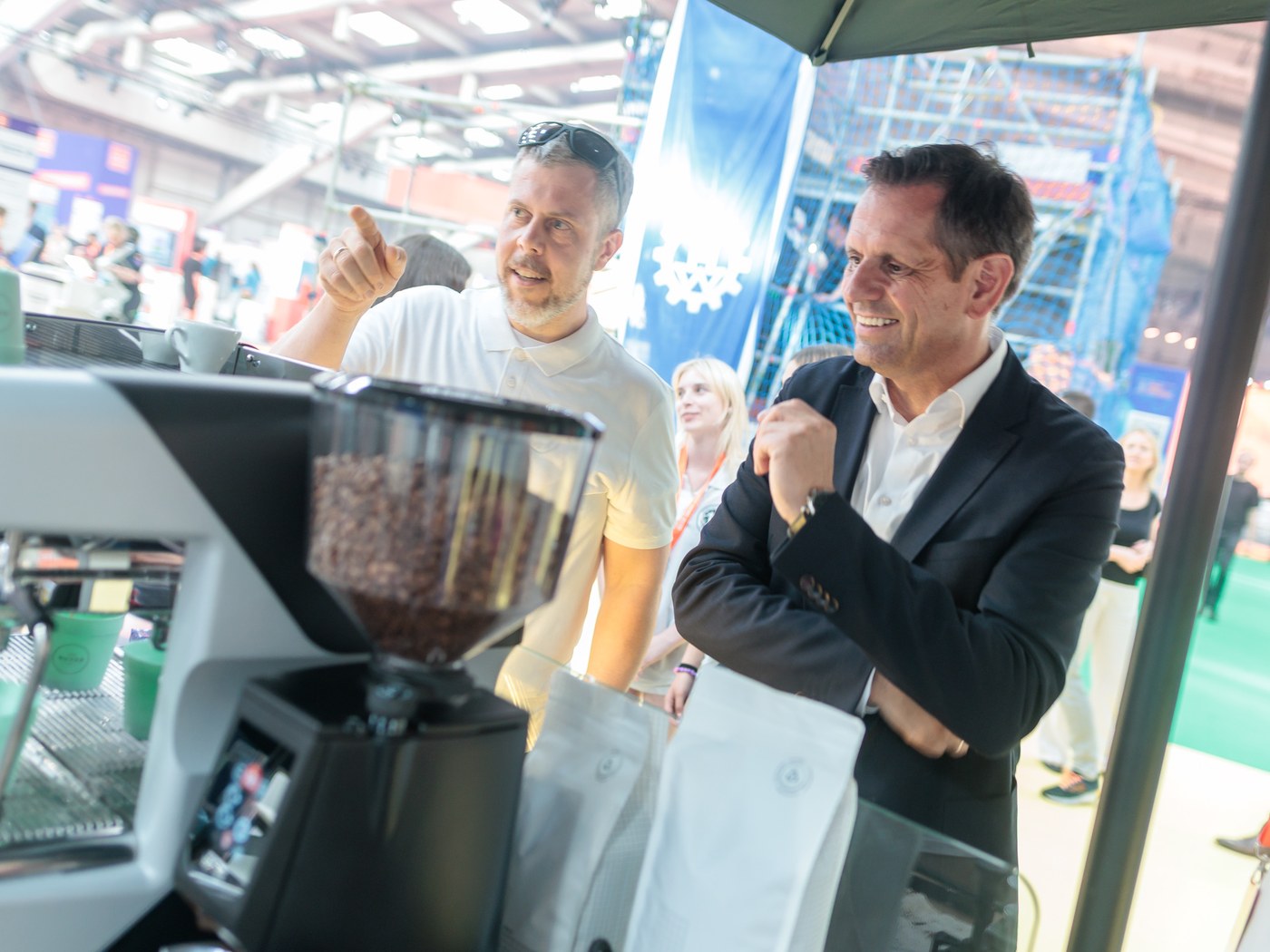 Helge Kletti erklärt einem Standbesucher an einer Kaffeemühle den Röstprozess, umgeben von Kaffeesäcken auf einer Messe