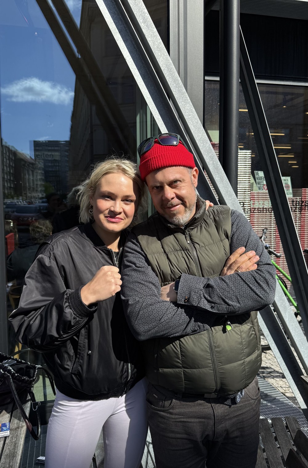 Helge Kletti und Clara-Sophia Müller posieren mit erhobener Faust und verschränkten Armen vor einer modernen Glasfassade