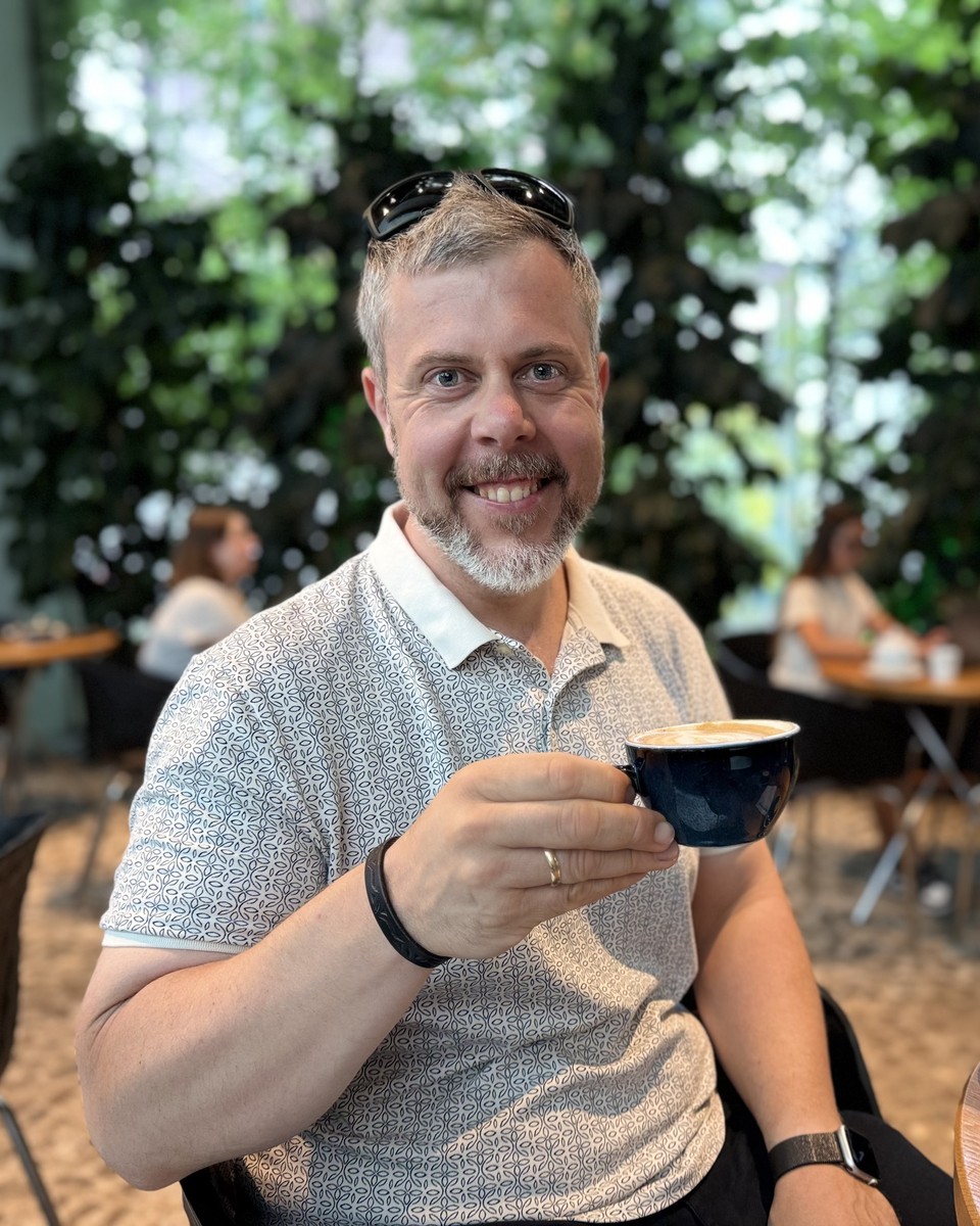 Helge Kletti lächelt in die Kamera und hält eine Tasse Kaffee, im Hintergrund ein begrüntes Café
