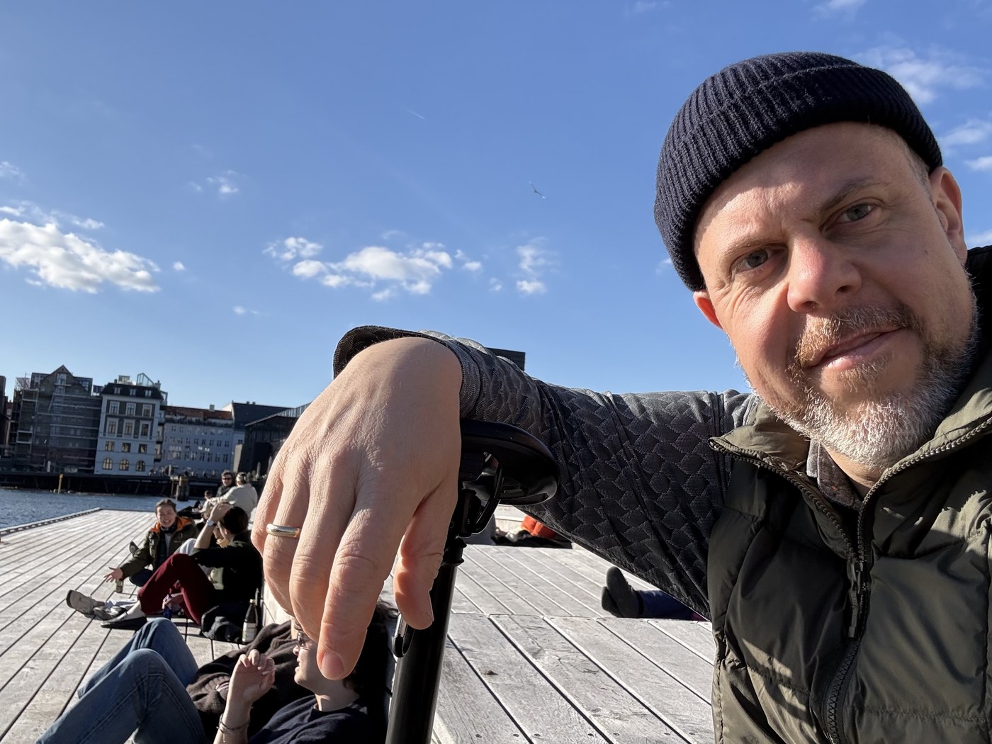 Helge Kletti im Nahporträt an einem Hafenkai, im Hintergrund Backsteinhäuser und blauer Himmel